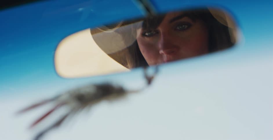 A woman's reflection in a car's rearview mirror as she drives with a feather charm hanging below