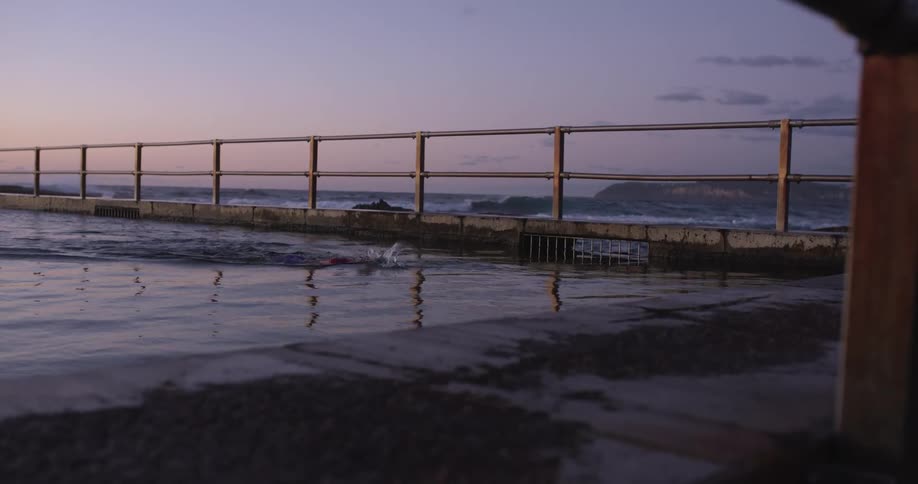 A lone swimmer does laps in a calm seaside rock pool during a beautiful sunrise as waves crash in the ocean behind