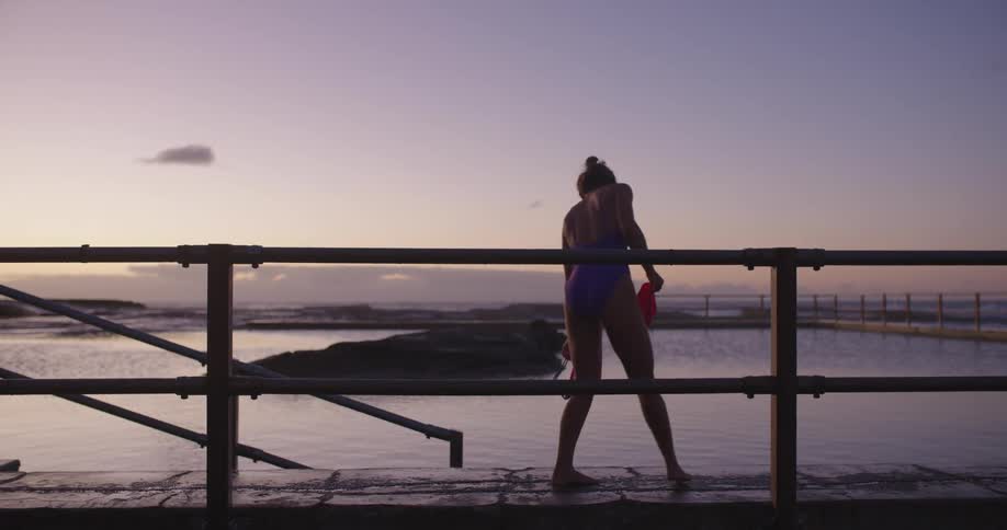 A female swimmer walks along the side of a tranquil ocean rock pool, preparing for her morning swim as the sun rises