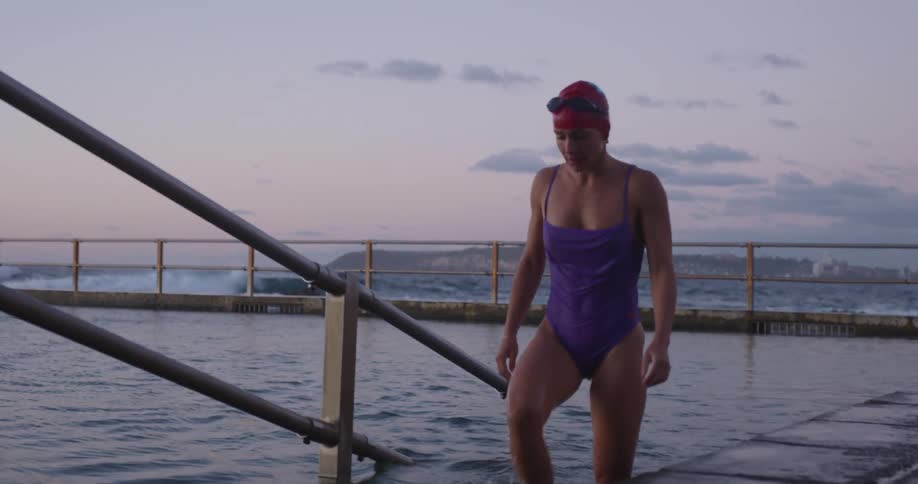 A woman in a purple swimsuit and red swim cap gets out of an ocean rock pool at dusk in slow motion