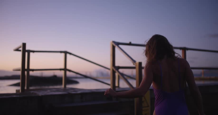 Following from behind, a female swimmer in a purple bathing suit walks in slow motion towards the water's edge at twilight