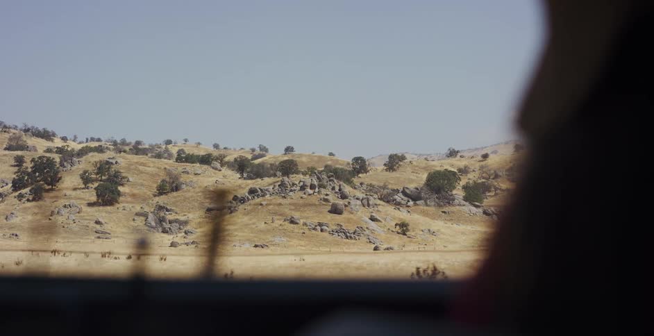 Scenic view of a dry, golden, and rocky hillside from the window of a moving vehicle on a sunny day