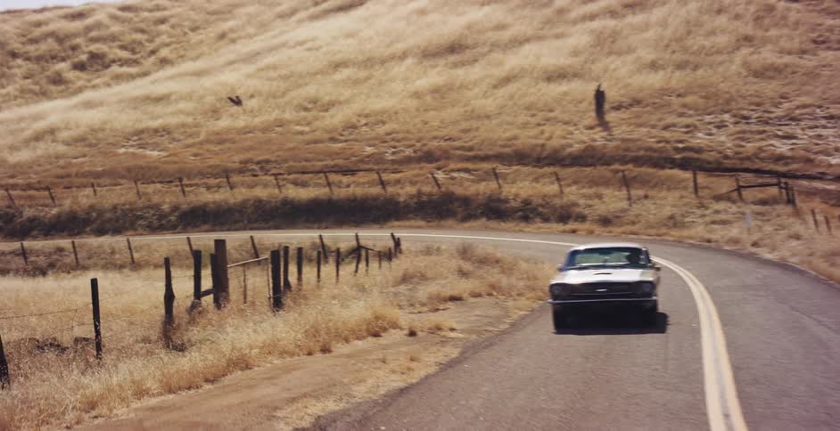 An aerial view follows a classic silver Ford Mustang as it drives along a winding country road through dry, golden hills