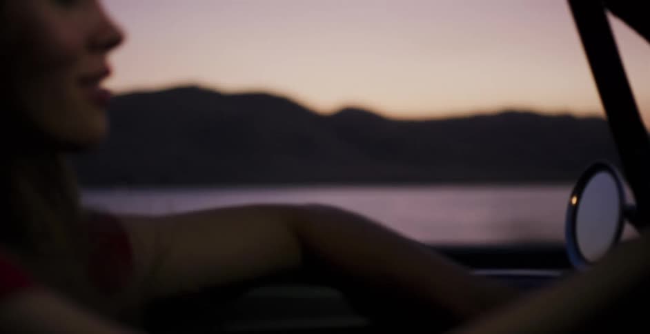 Close up on a smiling woman enjoying a carefree drive in a convertible along a scenic lakefront at dusk