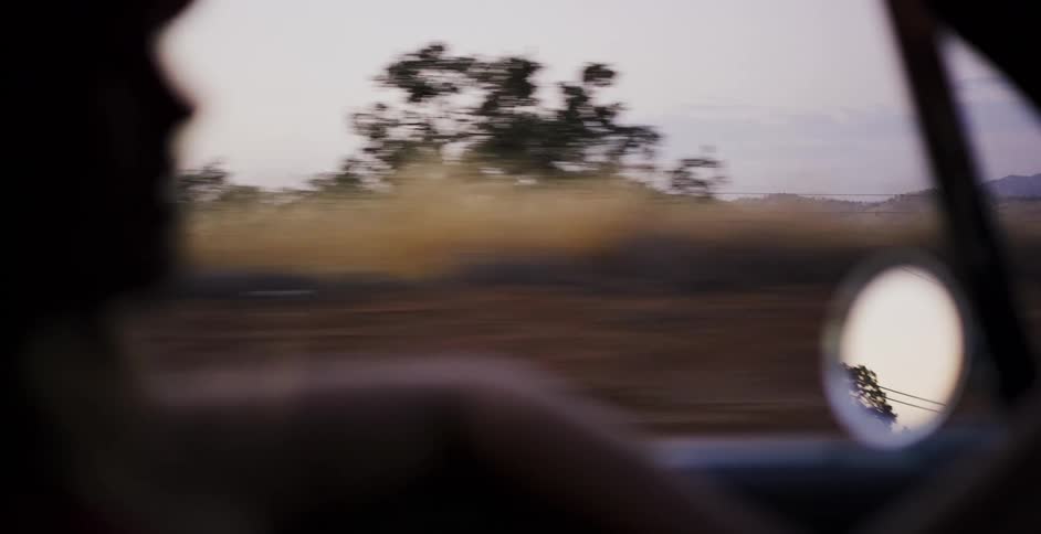 A passenger's view from inside a moving car reveals the passing arid, rural landscape with rolling golden hills at dusk