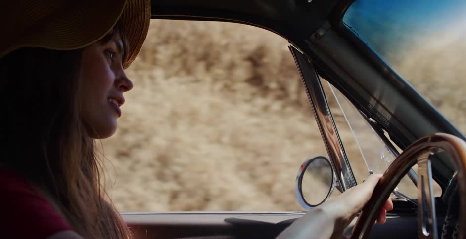 A stylish woman in a sun hat enjoys a drive in her vintage car along a scenic, sun-drenched road