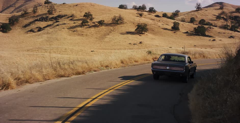 A classic muscle car drives down a winding country road through golden, rolling hills on a sunny day