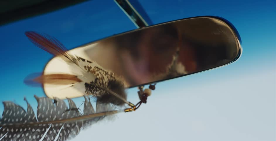 Decorative feathers hanging from a car's rearview mirror sway in the wind against a clear blue sky