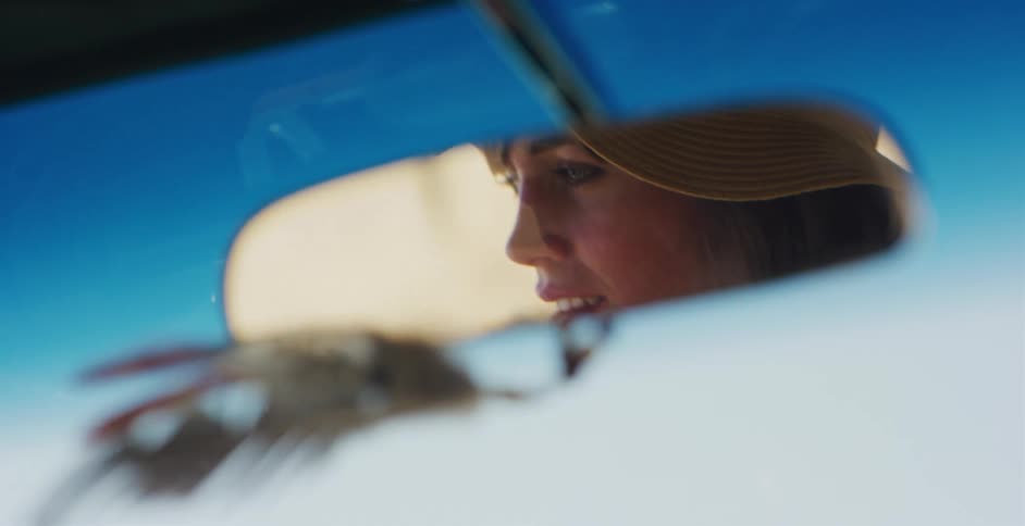 A slow-motion close-up shows the reflection of a woman in a sun hat driving her car as seen in the rearview mirror