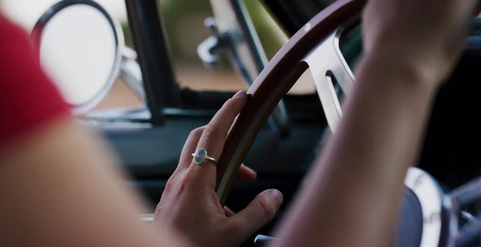 Close up from behind a woman in a red shirt driving a vintage car with her hands on the wooden steering wheel