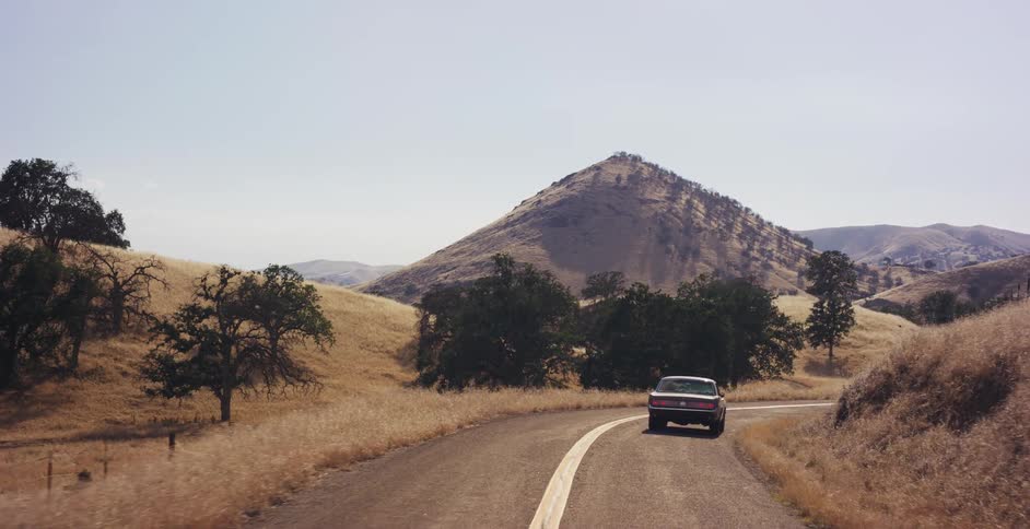 A classic vintage car takes a scenic drive along a winding road through the sun-drenched golden hills of the countryside