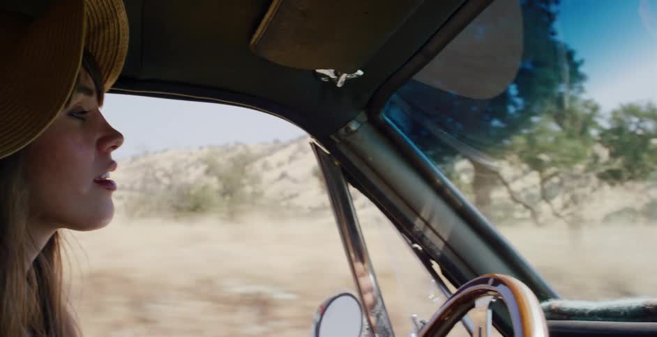 A woman in a yellow hat drives a vintage car down a dusty country road in slow motion
