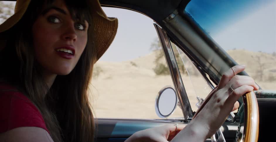A young woman wearing a straw hat happily drives a classic convertible through the sunny countryside in slow motion