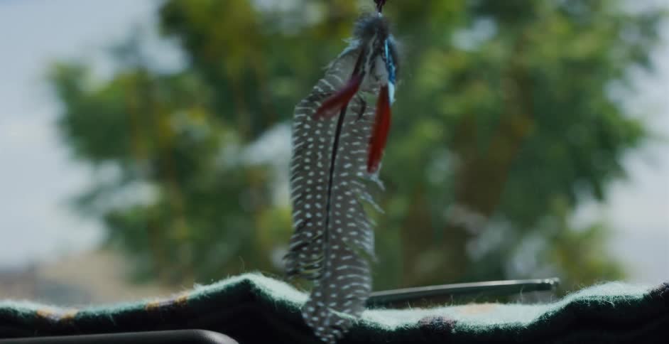 A close-up slow motion shot of a small bundle of natural feathers hanging from a car mirror and swaying gently against a blurred outdoor background