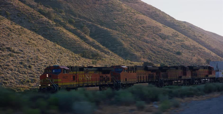An orange BNSF freight train travels along tracks through a rural mountain pass during golden hour