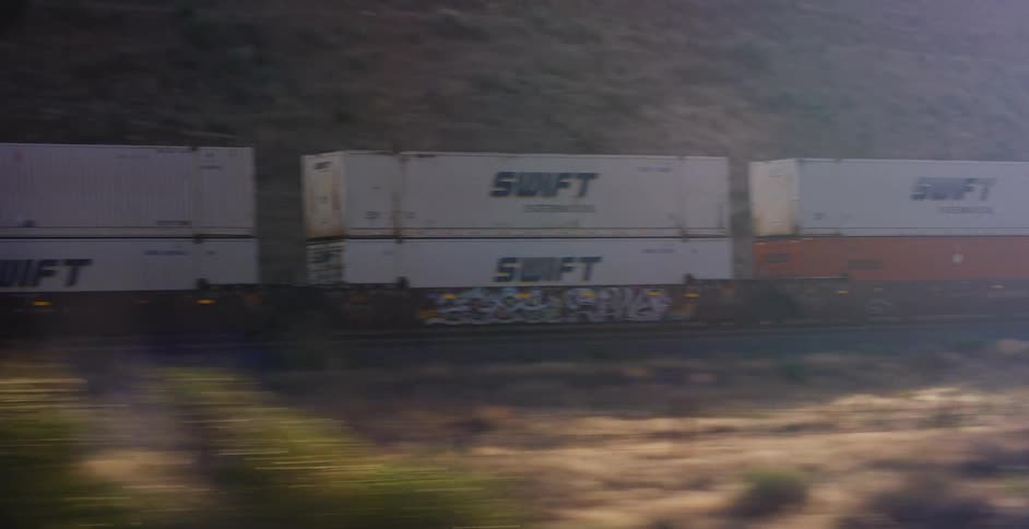 Side view of a fast-moving freight train with shipping containers traveling through an arid, mountainous region