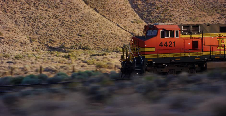 A BNSF locomotive travels along railroad tracks through a vast, arid landscape during a warm sunset