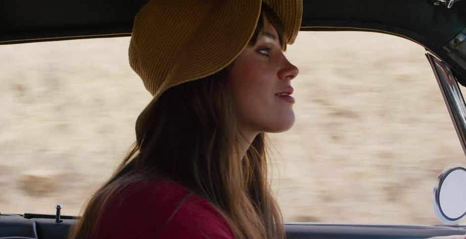 A beautiful woman in a floppy hat smiles while riding as a passenger in a car on a desert road trip