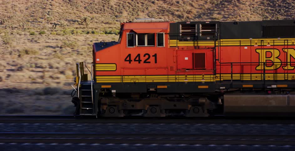 A close-up tracking shot of a BNSF freight train locomotive traveling through a desert landscape at golden hour