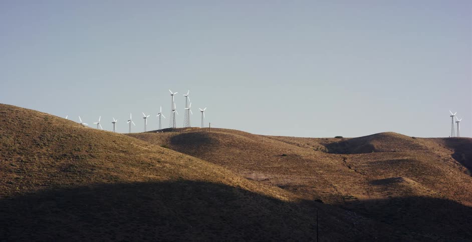 A wind farm with spinning turbines sits atop arid, rolling hills under a clear blue sky
