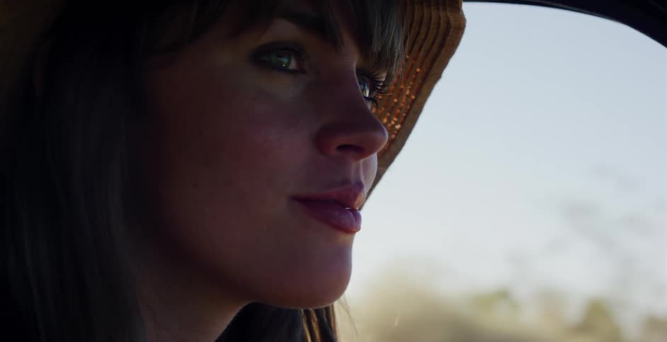 Close-up of a smiling woman in a straw hat driving through a desert landscape during golden hour