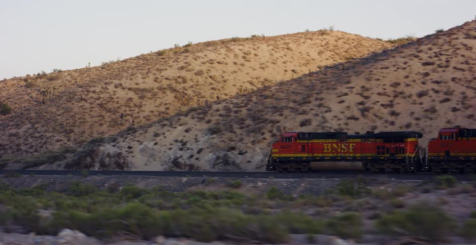A BNSF freight train travels along the tracks through a vast, hilly desert landscape at golden hour