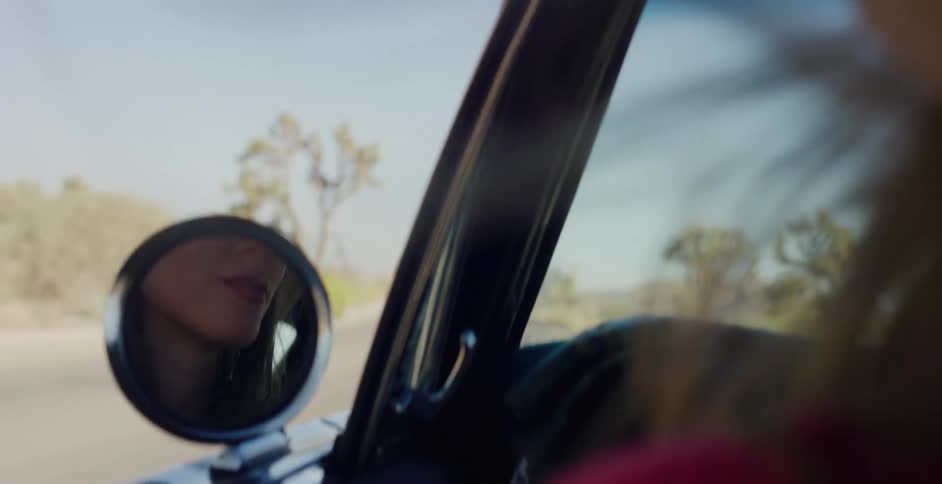 A woman's reflection is captured in the side view mirror of a vintage car as she drives down a desert highway on a sunny day
