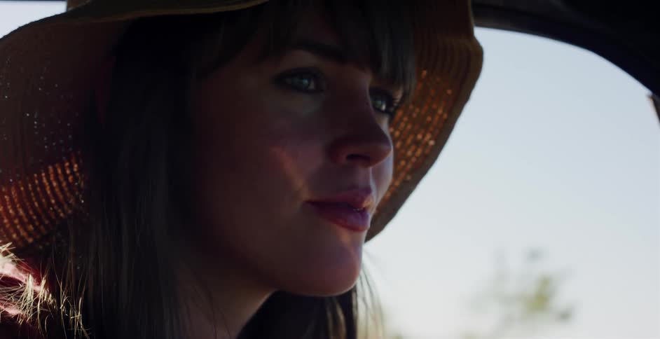 A sunlit close-up of a serene woman in a straw hat driving her car on a beautiful day