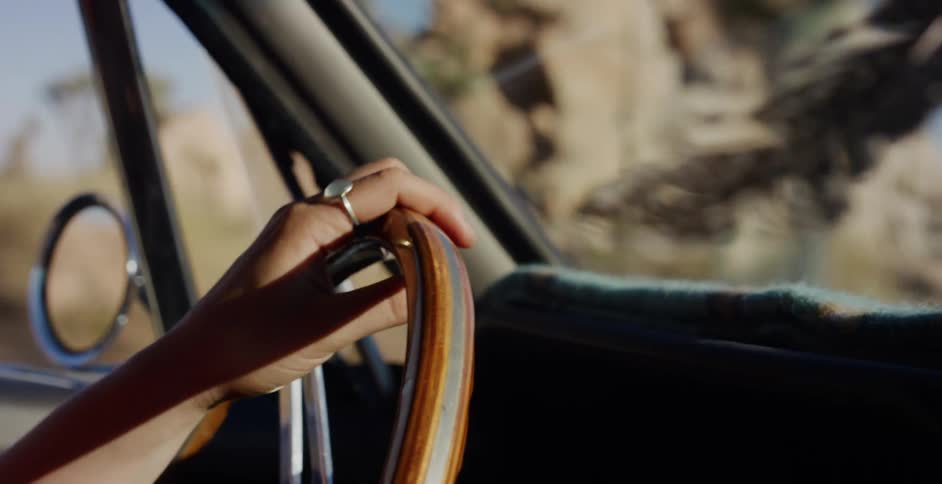 A woman's hand rests on a vintage wooden steering wheel as she drives through a sunny desert landscape on a road trip
