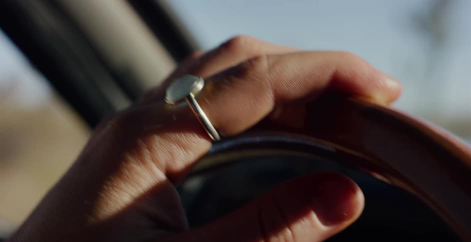 A close-up shot shows a hand wearing a silver ring resting on a wooden steering wheel while driving