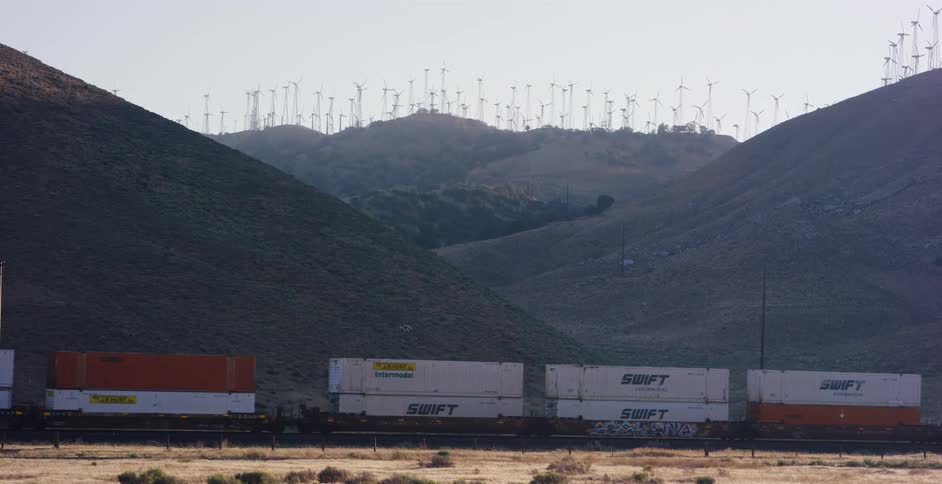 A freight train with shipping containers travels across a desert landscape in front of a massive wind farm on rolling hills