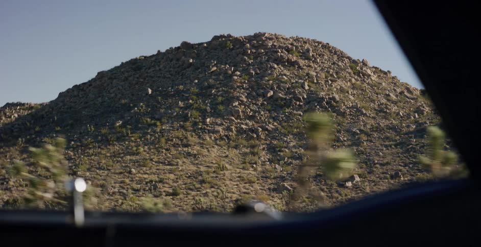 Passenger's point of view from a moving car shows a rocky desert mountain and Joshua trees passing by