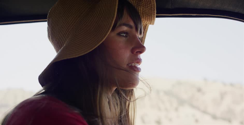 A beautiful woman in a floppy straw hat enjoys the wind blowing through her hair during a slow-motion car ride through a sunny landscape