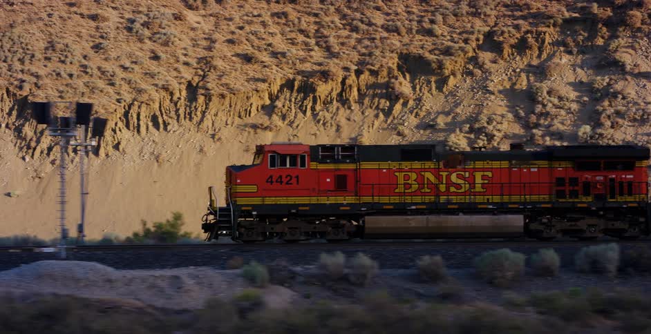 A side-view tracking shot follows a BNSF freight train moving through a desert landscape at golden hour