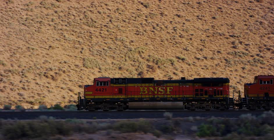 A BNSF freight train travels along the tracks through a vast, arid desert landscape during golden hour