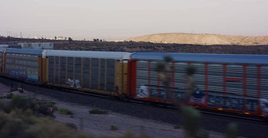 An aerial view tracks a long freight train with graffiti traveling around a bend in the Mojave Desert at twilight