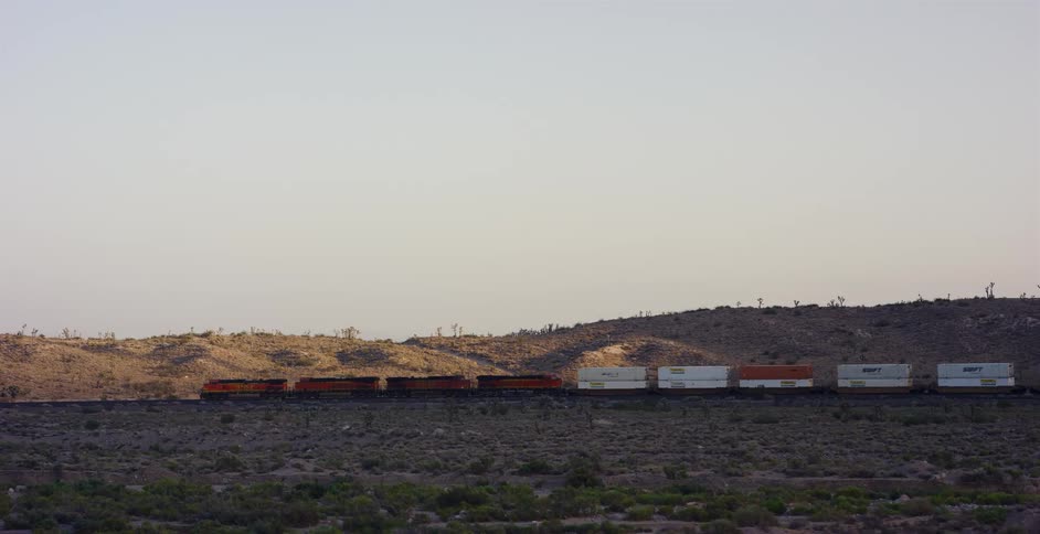 A long freight train carries shipping containers as it travels through the vast, arid desert landscape