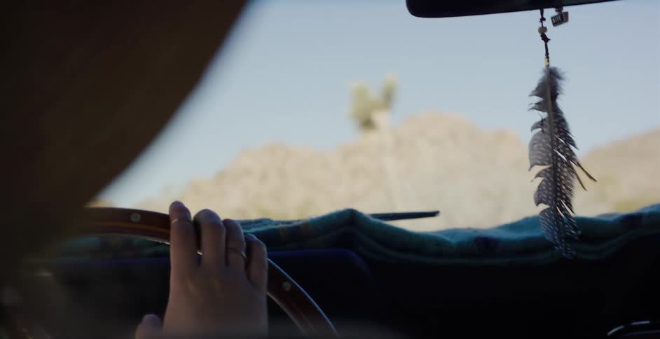 A feather sways from the rearview mirror of a car as a woman drives down a scenic desert road on a sunny day