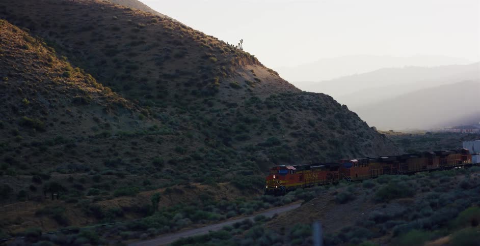 An aerial tracking shot follows a long freight train as it travels through a hazy, sunlit mountain valley