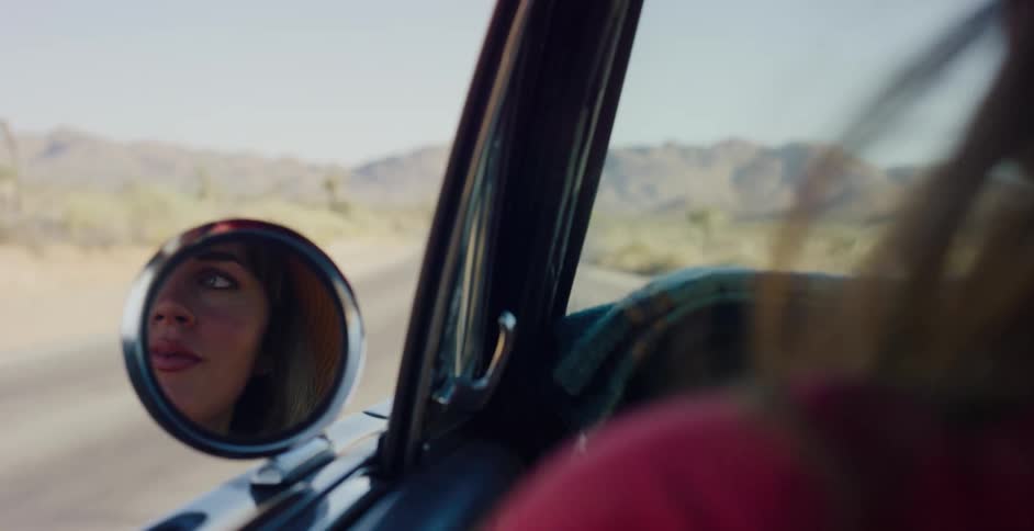 A smiling woman's reflection in the side mirror of a vintage car as she drives through a sunny desert landscape