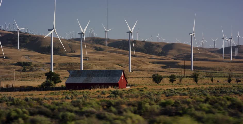 A slow-motion tracking shot passes a classic red barn nestled amongst a vast wind farm on rolling golden hills