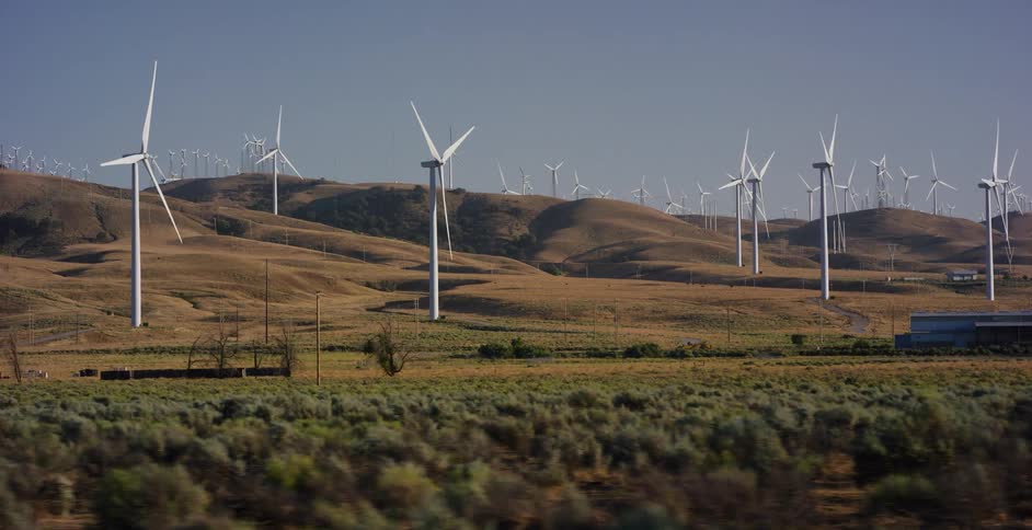 A slow-motion tracking shot reveals a vast wind farm with dozens of spinning turbines spread across the dry, rolling hills