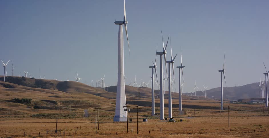 A cinematic tracking shot reveals a vast wind farm with numerous turbines generating renewable energy across the arid, golden hills of California