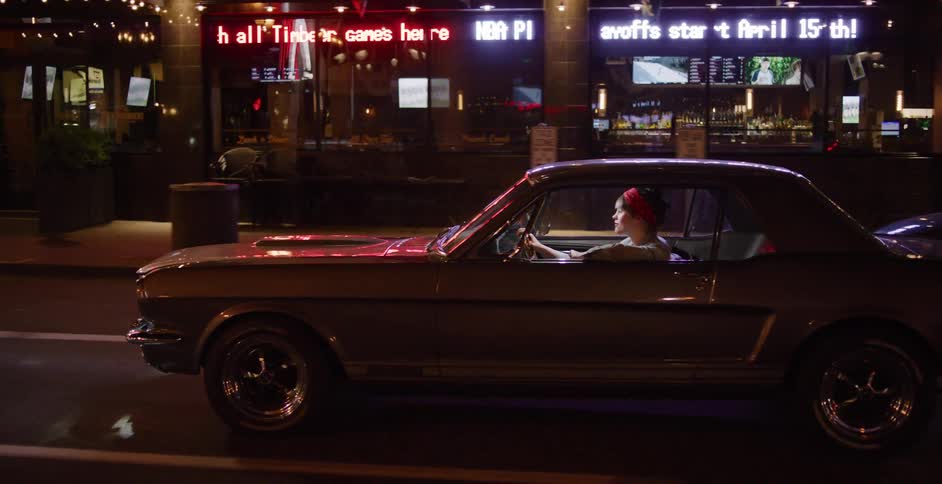 A stylish woman drives a classic Ford Mustang down a city street illuminated by neon lights at night