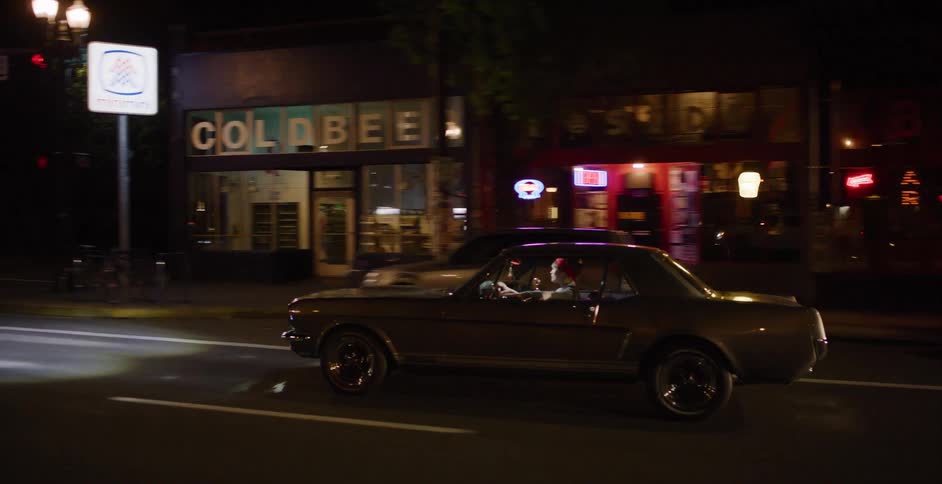 Side profile tracking shot of a classic silver Ford Mustang driving through a vibrant city at night