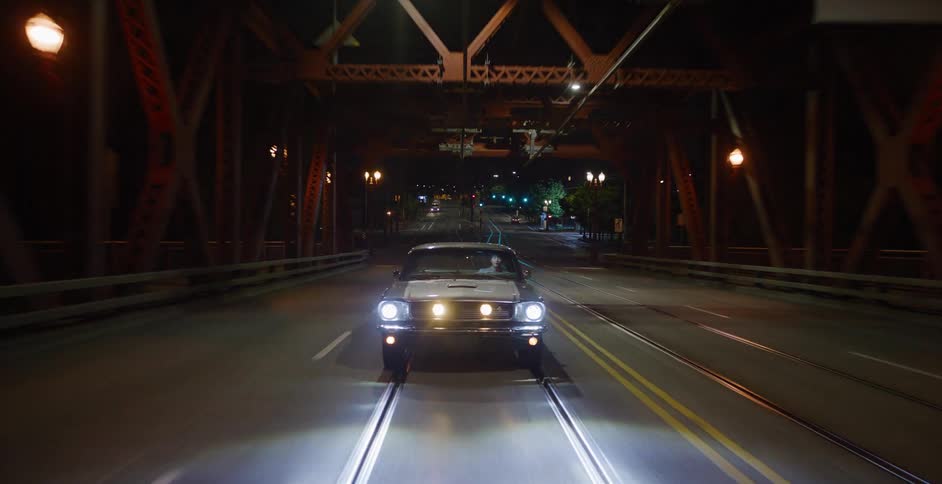 A classic muscle car with bright headlights drives across an empty city bridge at night