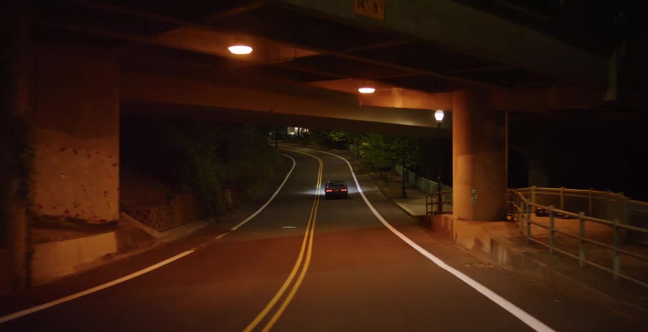 Following a car down a winding city road at night, passing under an illuminated overpass