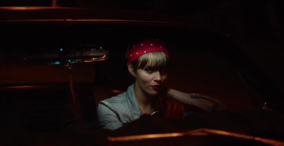 A stylish woman in a retro car waits on a city street at night, illuminated by passing lights