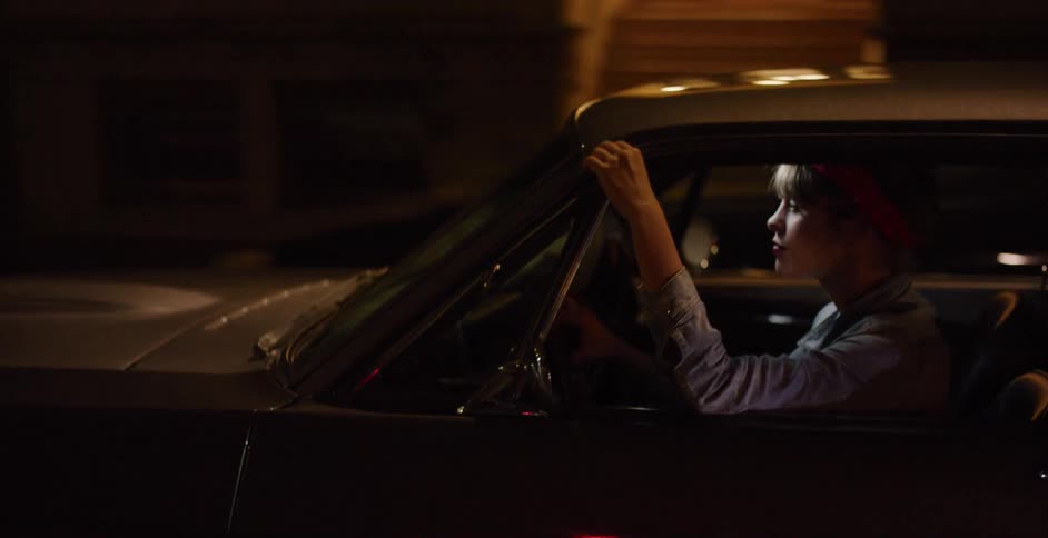 A cool young woman with a red bandana drives her classic muscle car through the city streets at night