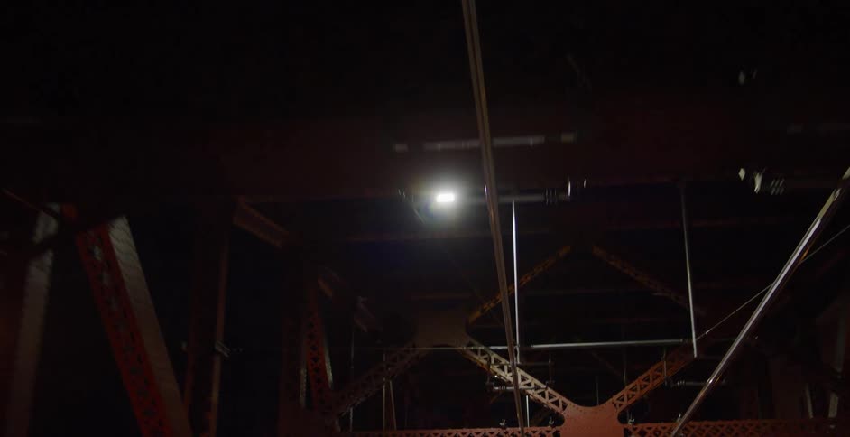 A slow, upward-looking point-of-view shot of driving under the illuminated steel truss structure of a bridge at night